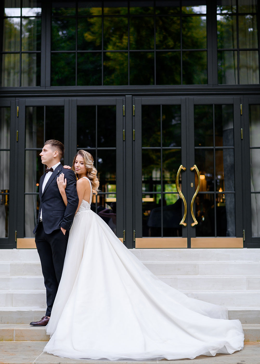 Side view of confident and attractive groom in stylish suit, keeping hands in pocket, standing and looking forward while his  stunning wife, wearing in long elegant silk dress, hugging him shoulders while posing outdoor on background of large transparent black doors of contemporary building